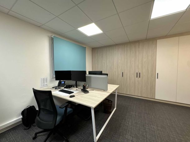 Bureau professionnel aménagé sur mesure avec une grande armoire en bois clair et blanc, un bureau en bois clair équipé d'écrans doubles, et un store bleu apportant une touche de couleur à un espace de travail moderne.