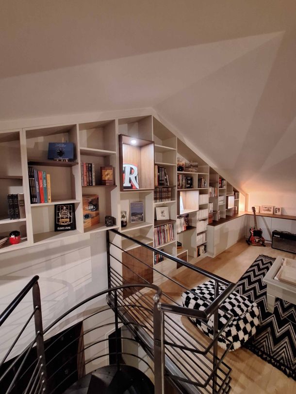 Bibliothèque sur mesure sous pente avec étagères ouvertes et niches éclairées, située dans un espace mezzanine, accompagnée d'un escalier en colimaçon avec garde-corps en métal noir, parquet clair, et décoration cosy avec tapis graphique et guitare électrique en arrière-plan.