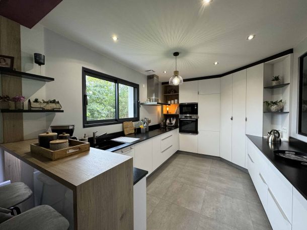 Cuisine contemporaine spacieuse avec des façades blanches, un plan de travail noir, des étagères ouvertes et un îlot en bois, éclairée naturellement par une grande fenêtre avec vue sur l'extérieur.