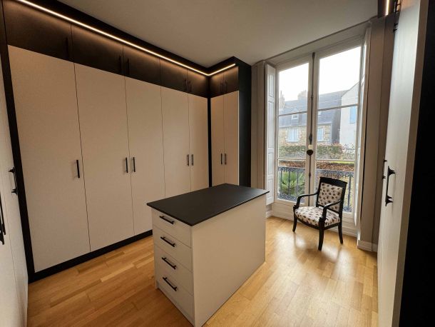 Dressing élégant avec armoires blanches et noires sur toute la hauteur, îlot central avec plateau noir, parquet en bois clair, éclairage LED intégré et une grande fenêtre laissant entrer la lumière naturelle